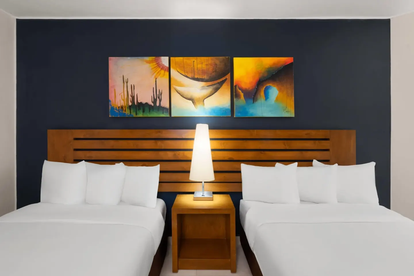 Standard Room with two beds, central lamp, wood headboard, and wall art at Grand Decameron Los Cabos.