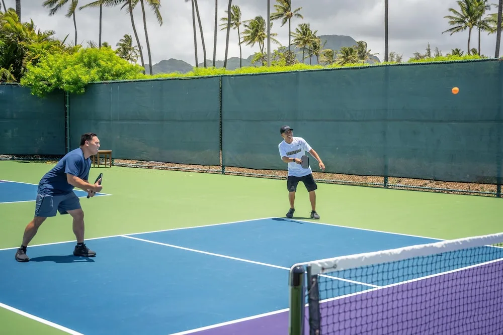 Pickleball players on an outdoor court at Grand Hyatt Kauai Resort and Spa and Spa with palms beyond the fence.