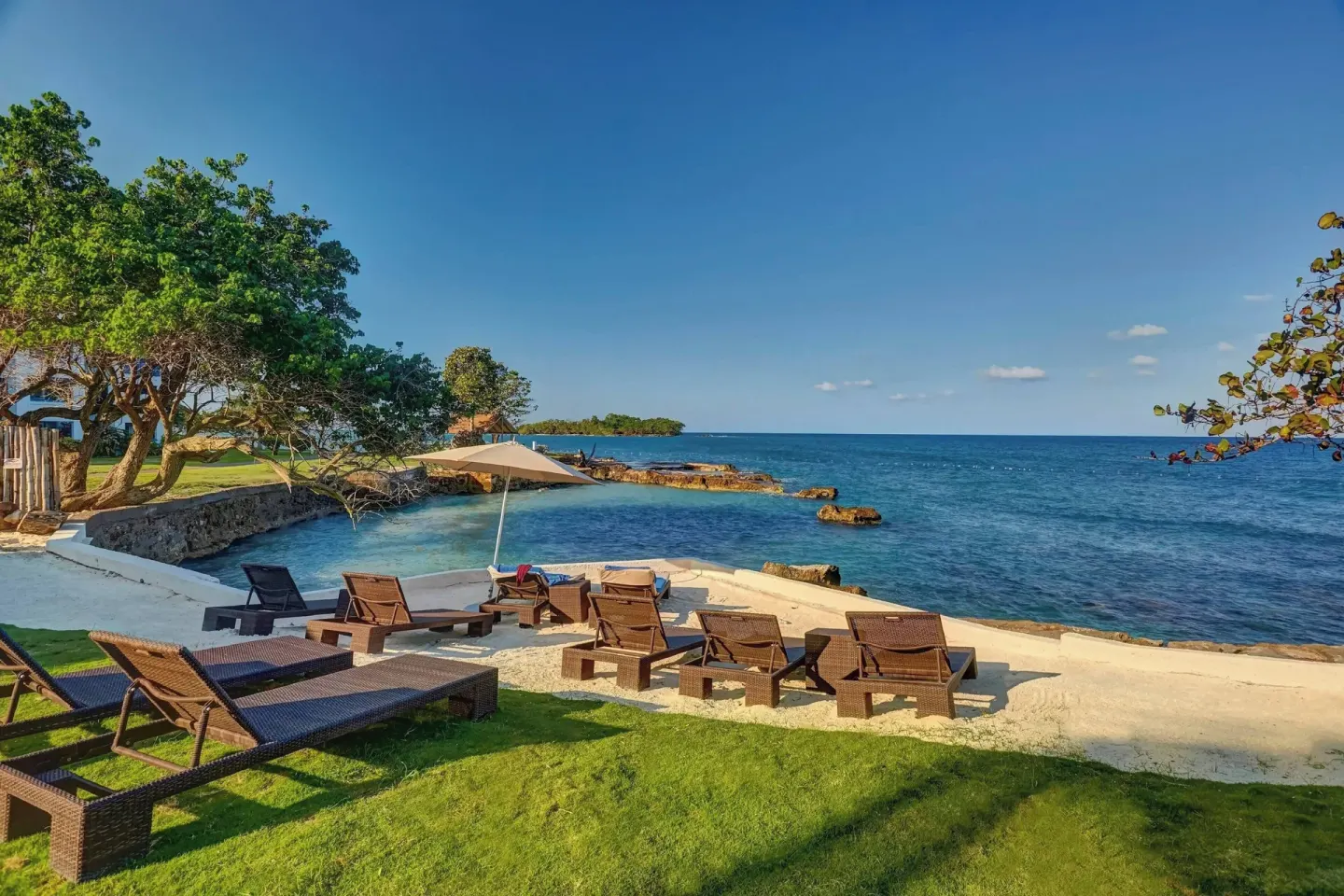 Oceanfront lounge chairs at Grand Lido Negril Au-Naturel set on grass and sand by rocky shoreline.