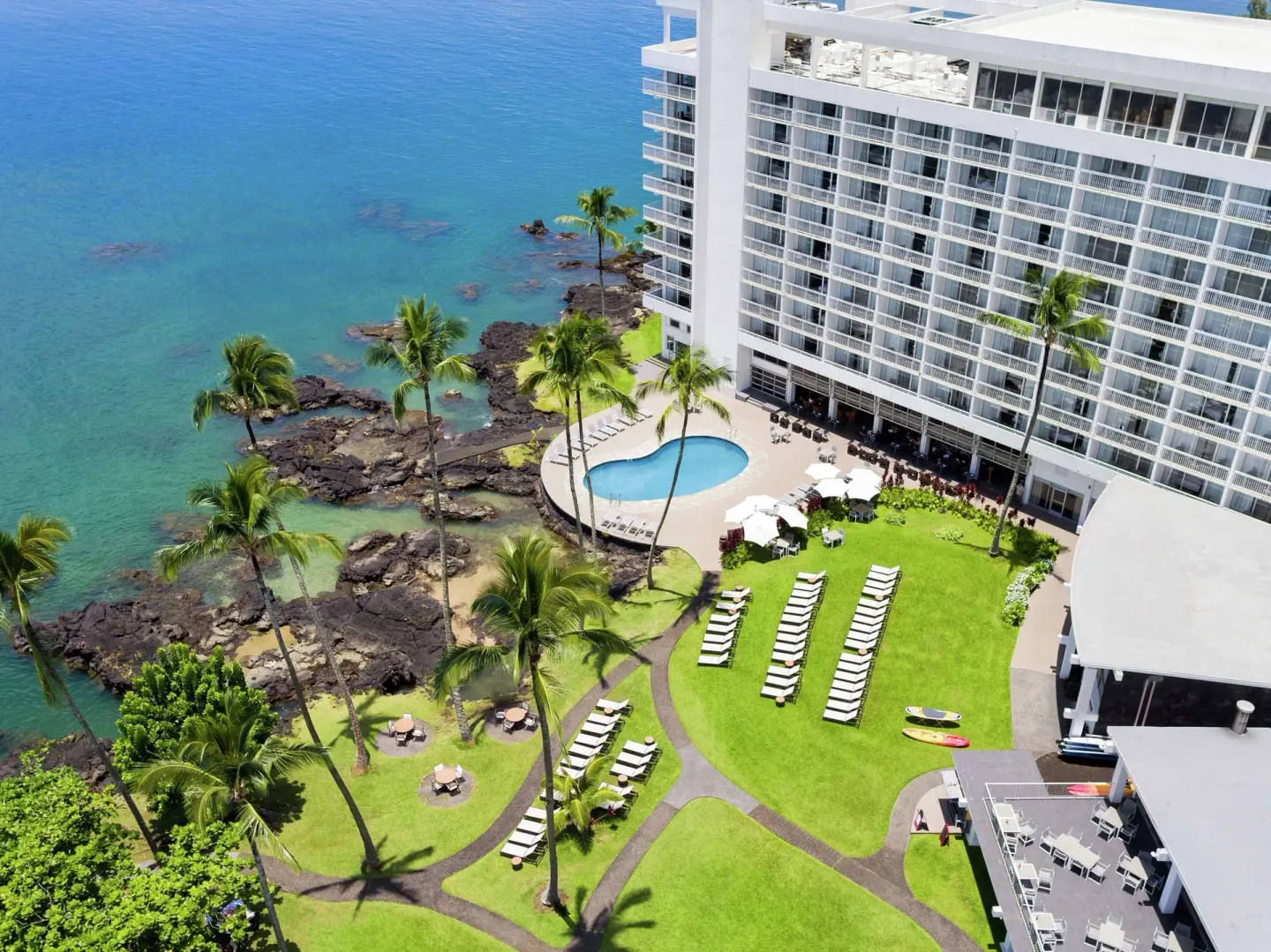 Aerial pool lawn at Grand Naniloa Hotel Hilo with palms, lounge chairs, lava shoreline, and hotel tower.