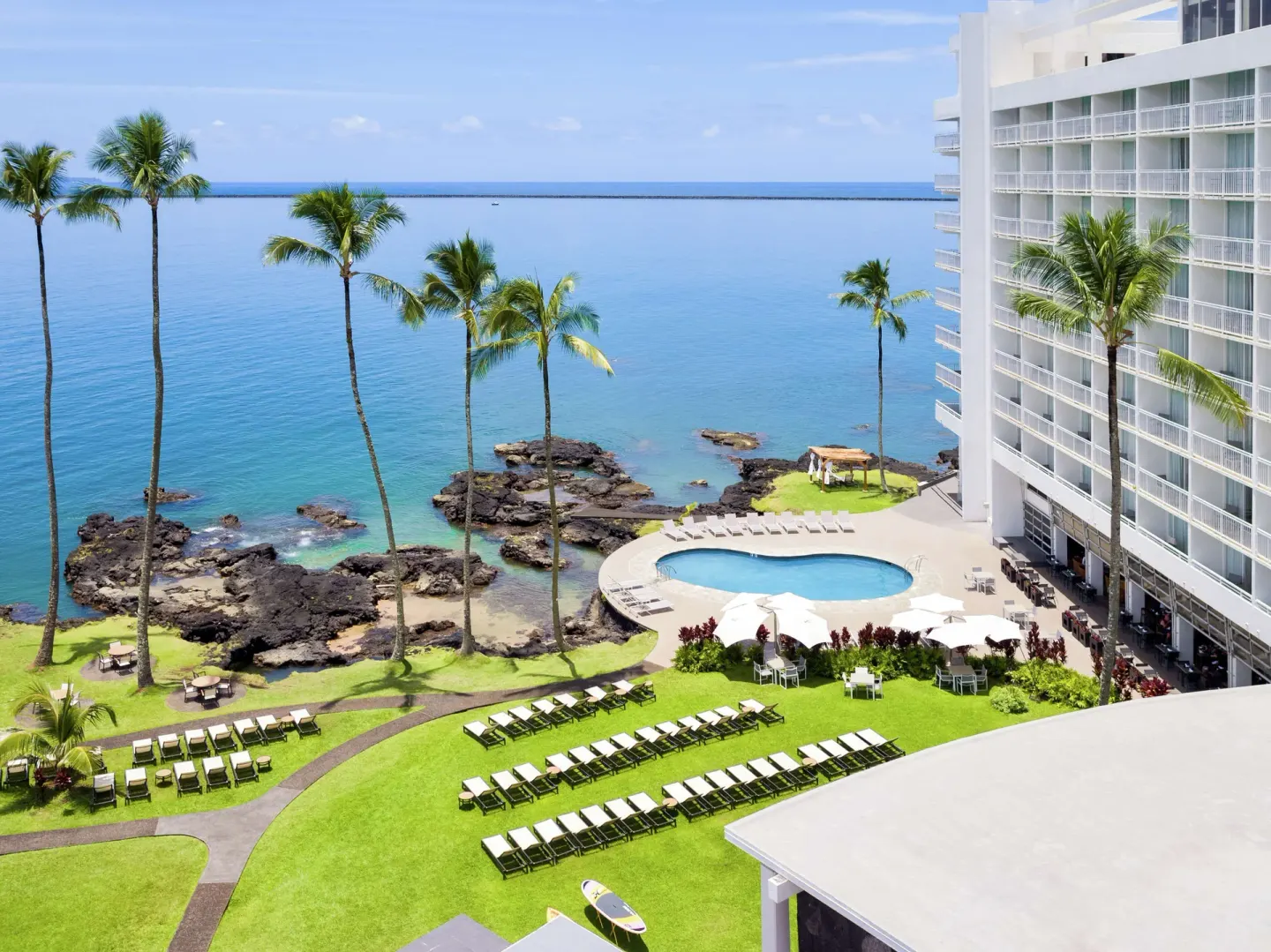 Pool and oceanfront lawn at Grand Naniloa Hotel Hilo with lounge chairs, palms, lava rocks, and bay views.