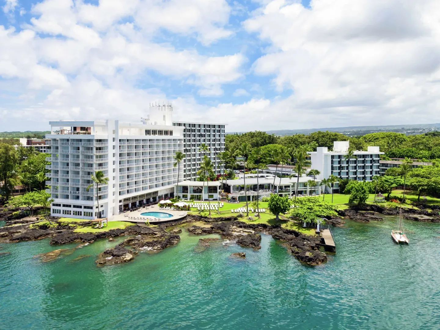Aerial view of Grand Naniloa Hotel Hilo on Hilo Bay with oceanfront lawns, pool, palms, and lava rocks.