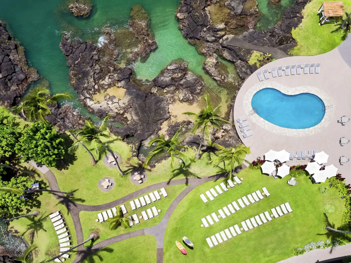 Overhead view of Grand Naniloa Hotel Hilo pool, lawns, loungers, palms, lava rocks, and turquoise water.