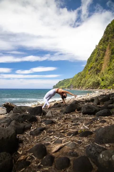 A person does a backbend on a rocky coast near Grand Naniloa Hotel Hilo.