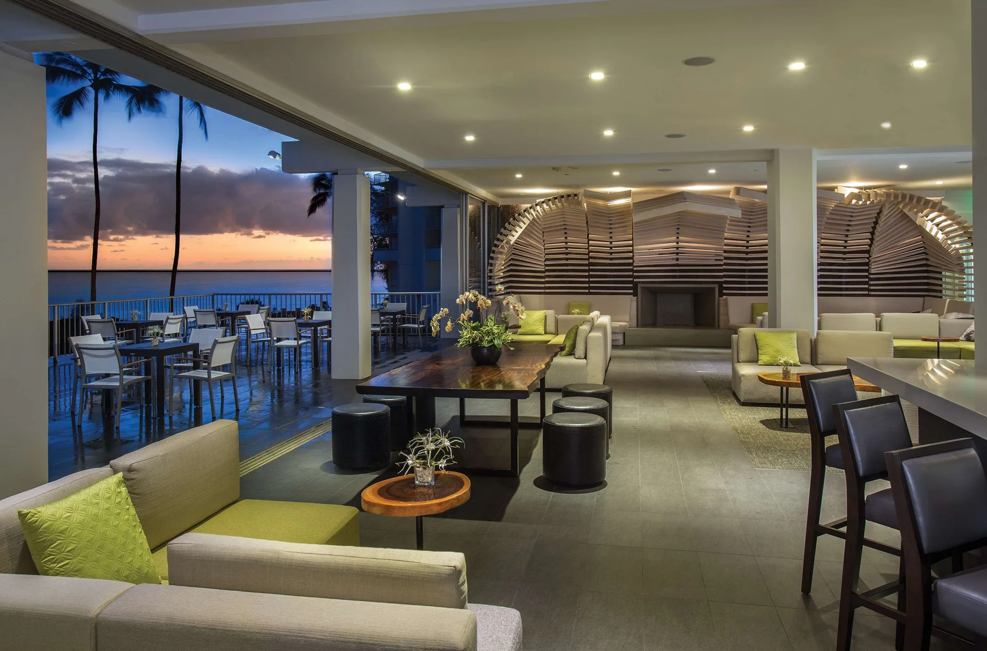 Open-air lounge seating at Grand Naniloa Hotel Hilo with bar chairs and an ocean sunset beyond the terrace.