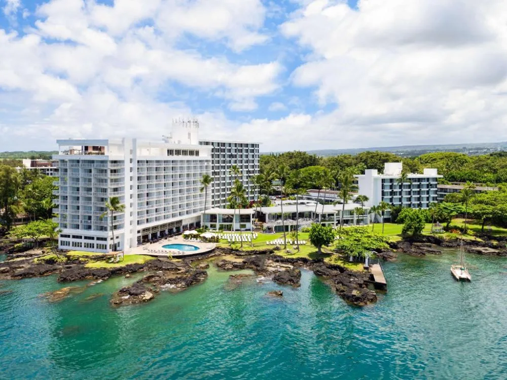 Oceanfront exterior of Grand Naniloa Hotel Hilo with bay, pool, lawns, and rocky shoreline.