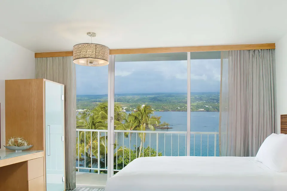Oceanfront lanai guest room with bed and bay view at Grand Naniloa Hotel Hilo.