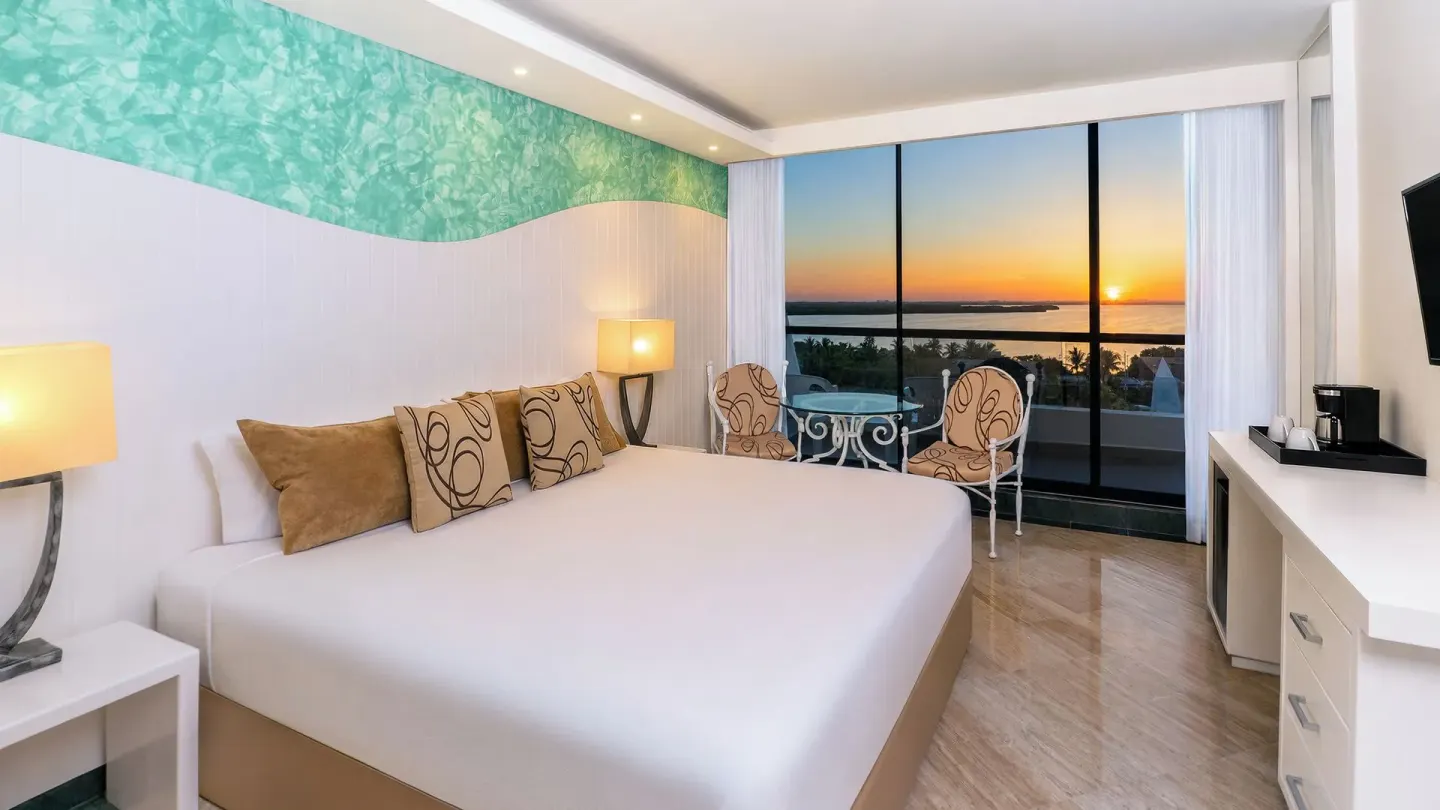 Grand Sunset View king room at Grand Oasis Cancun with bed, seating, balcony doors, and sunset water view.