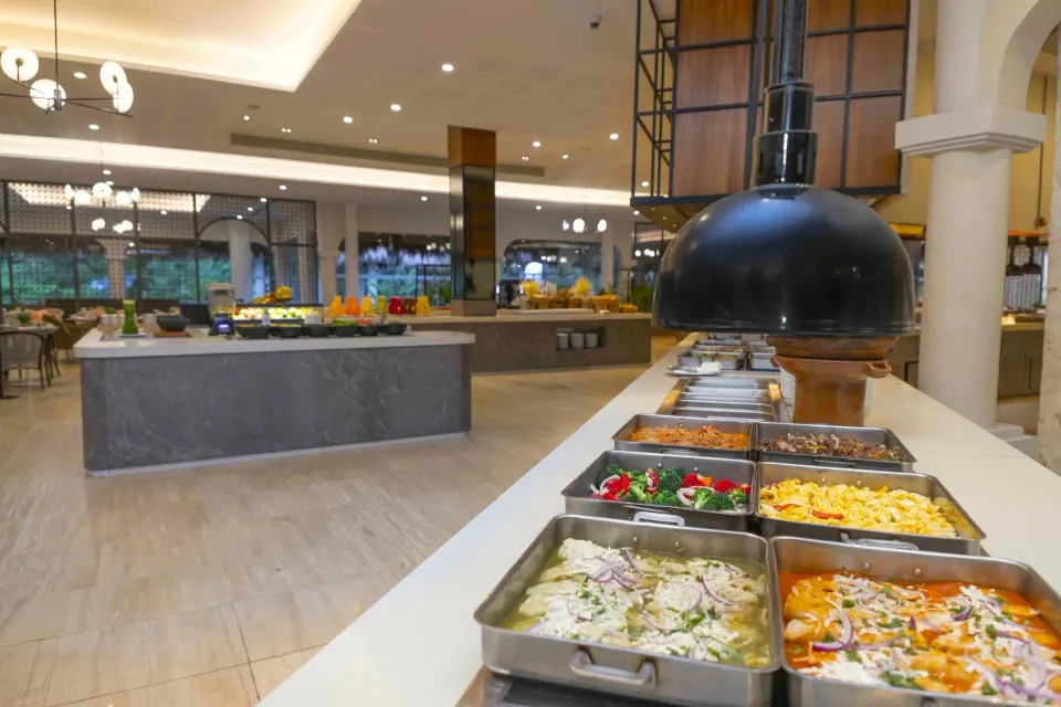 Buffet food trays on a long counter with the dining room beyond at Grand Palladium Colonial Resort & Spa