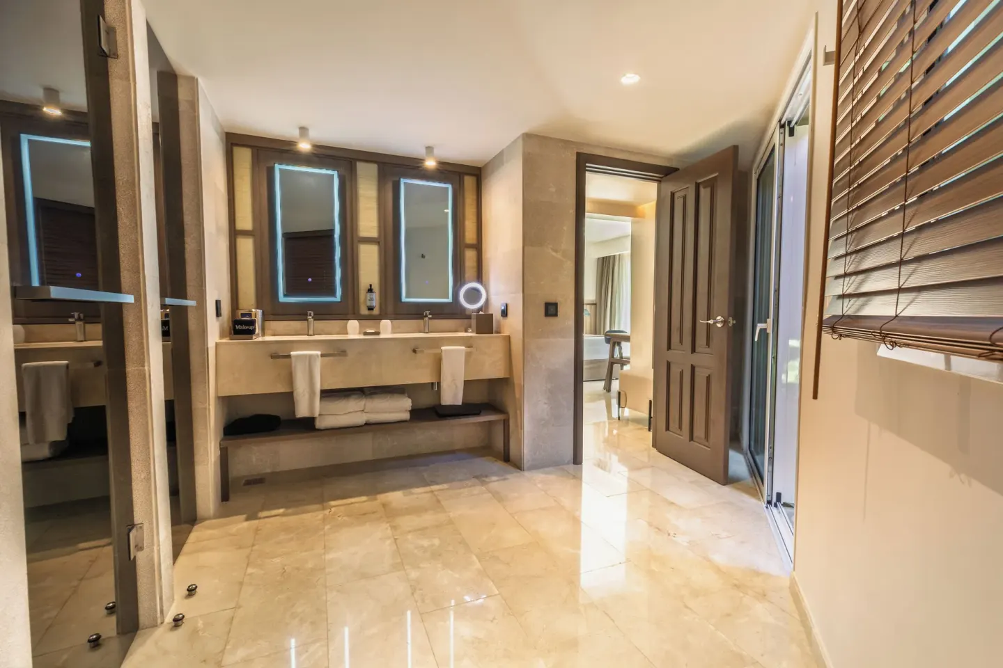 Guest bathroom with twin sinks, glass shower doors, and wooden blinds at Grand Palladium Select Bávaro.