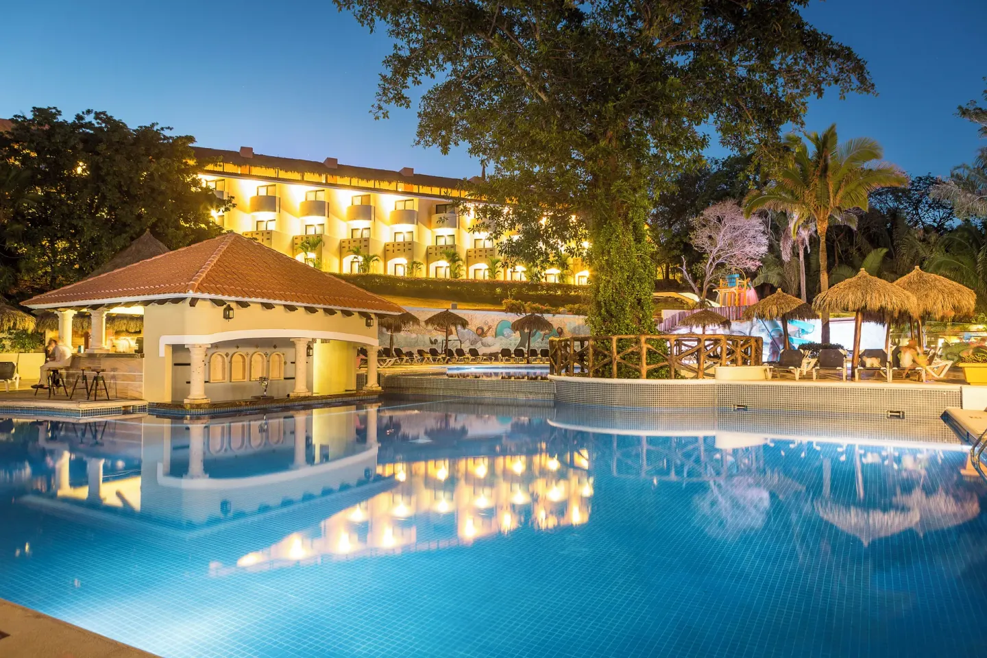 Evening pool scene with lit buildings and palapas at Grand Palladium Vallarta Resort & Spa.