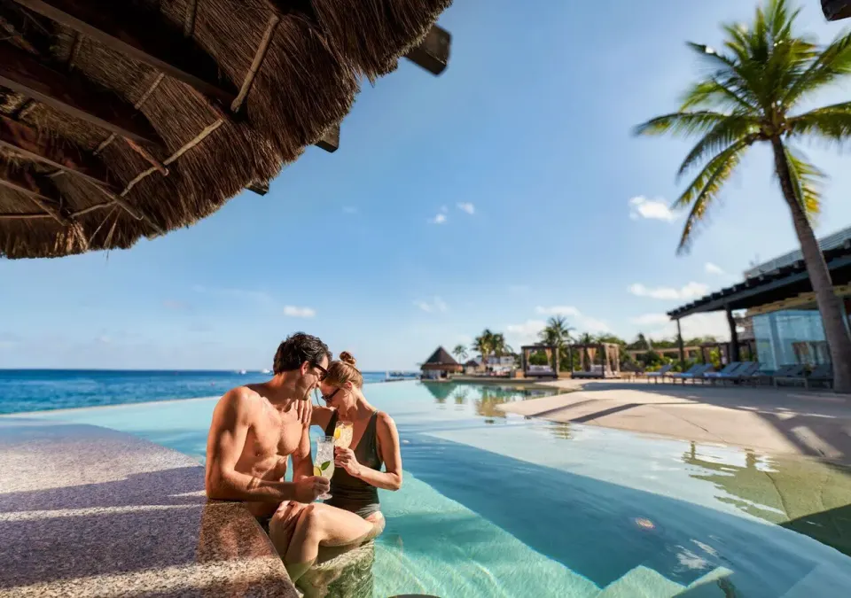 Couple sitting at the edge of a resort pool under a palapa at Grand Park Royal Cozumel.