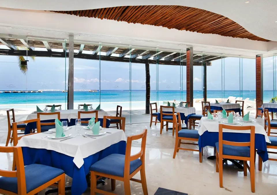 Seaside dining room with tables set and floor‑to‑ceiling windows at Grand Park Royal Cozumel