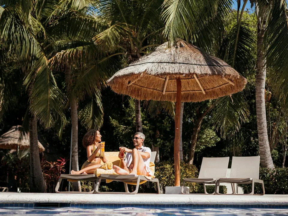Couple sitting on loungers under a palapa by the pool at Grand Sirenis Riviera Maya Resort & Spa