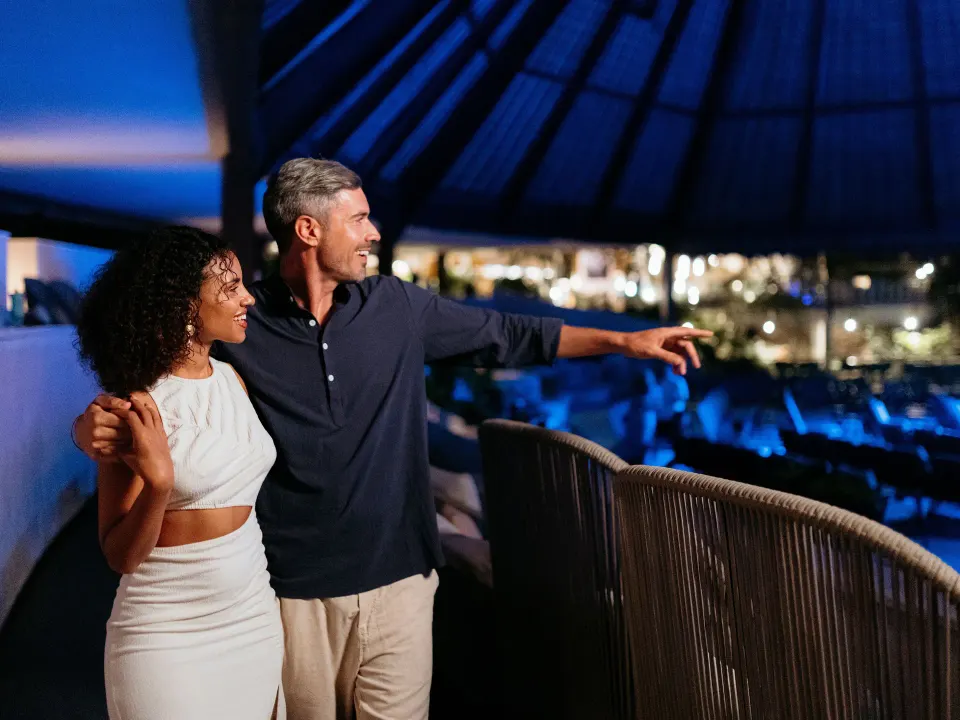 Couple standing on an elevated palapa balcony overlooking the resort at night at Grand Sirenis Riviera Maya Resort & Spa