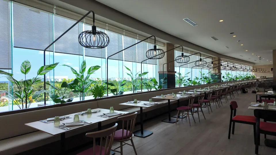 Long windowed dining room with plants and pendant lights at Grand Sirenis Riviera Maya Resort & Spa