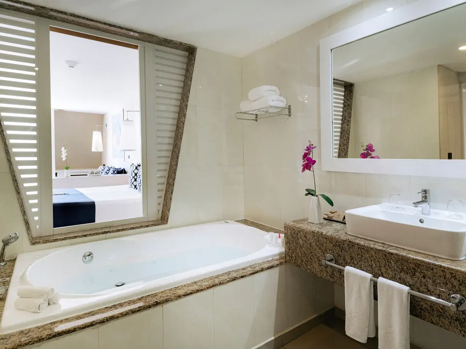 Bathroom whirlpool tub and sink with shutter window looking into bedroom at Grand Sirenis Riviera Maya Resort & Spa