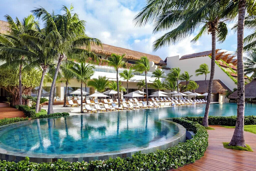 Curved pool, palms and rows of loungers beside resort buildings at Grand Velas Riviera Maya.