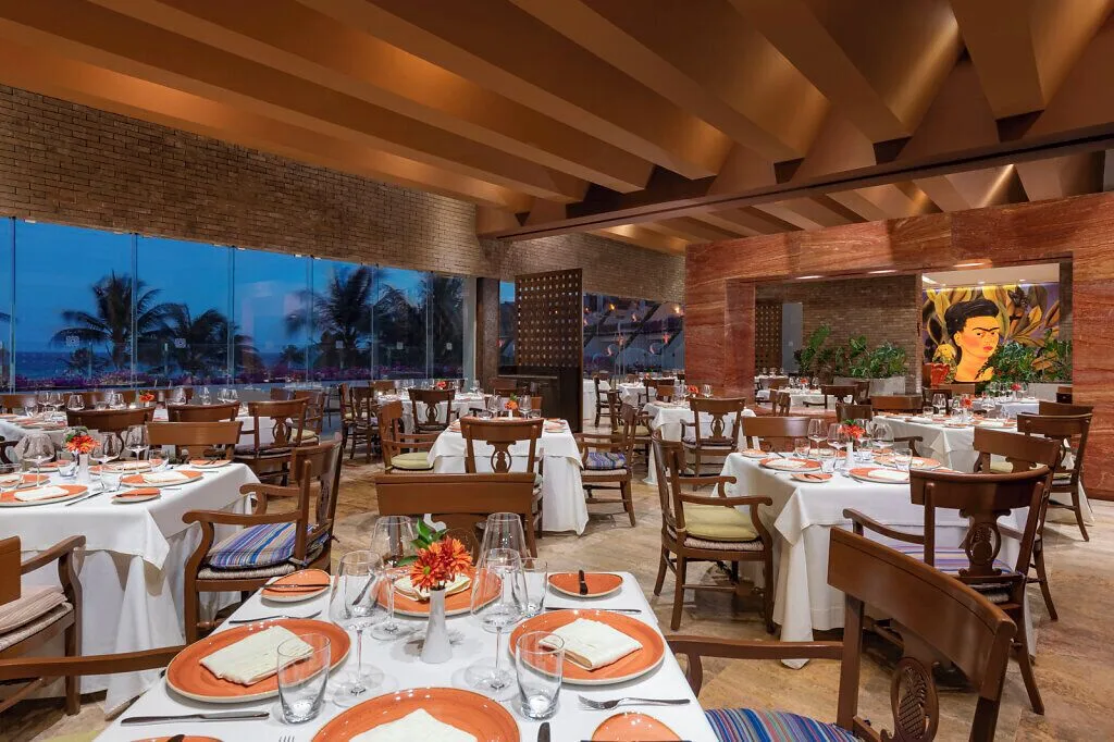 Frida restaurant at Grand Velas Riviera Maya with white tablecloths, orange plates, and portrait artwork.