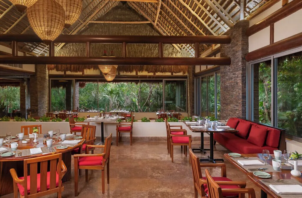 Garden-view restaurant at Grand Velas Riviera Maya with red cushions, wood tables, and a thatched roof.