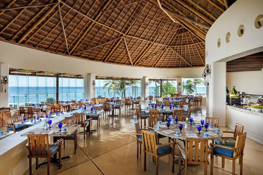 Oceanfront dining room at Grand Velas Riviera Maya with blue glassware and a thatched ceiling.