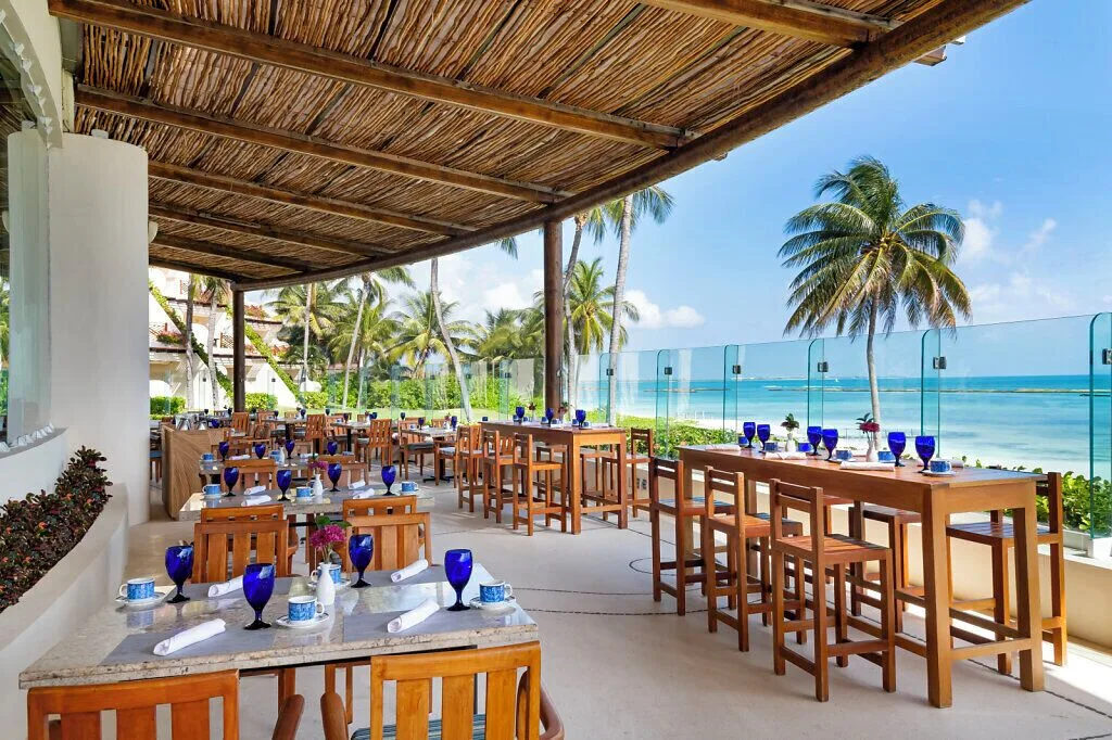 Seaside terrace dining at Grand Velas Riviera Maya with wooden tables, blue glasses, and palm views.