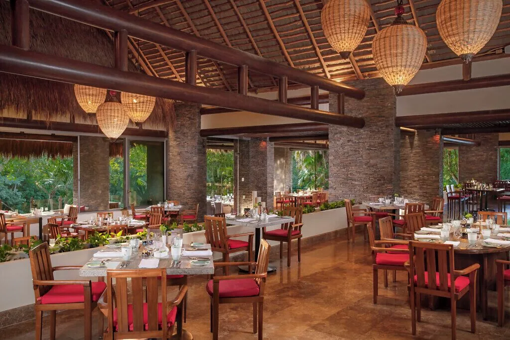 Thatched-roof restaurant at Grand Velas Riviera Maya with red-cushioned chairs and woven pendant lights.