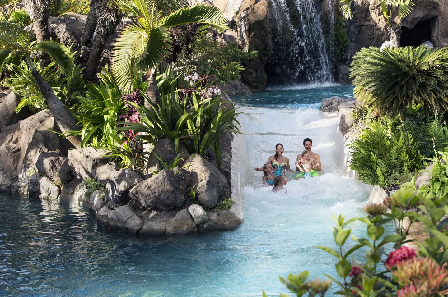 Family riding a white pool slide beside waterfalls and tropical rocks at Grand Wailea Maui.