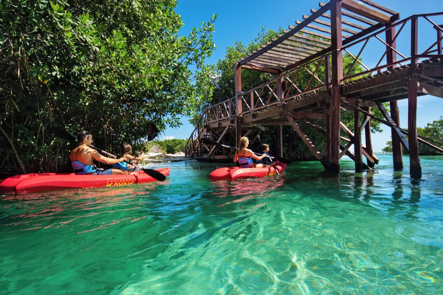 Guests kayaking on clear water beside a wooden bridge at Hacienda Tres Rios Resort, Spa & Nature Park.
