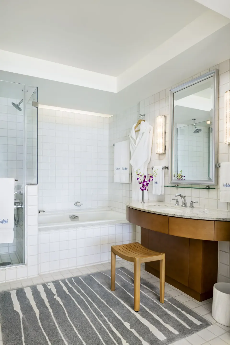 Bathroom with soaking tub, glass shower, vanity, robe, and striped rug at Halekulani.