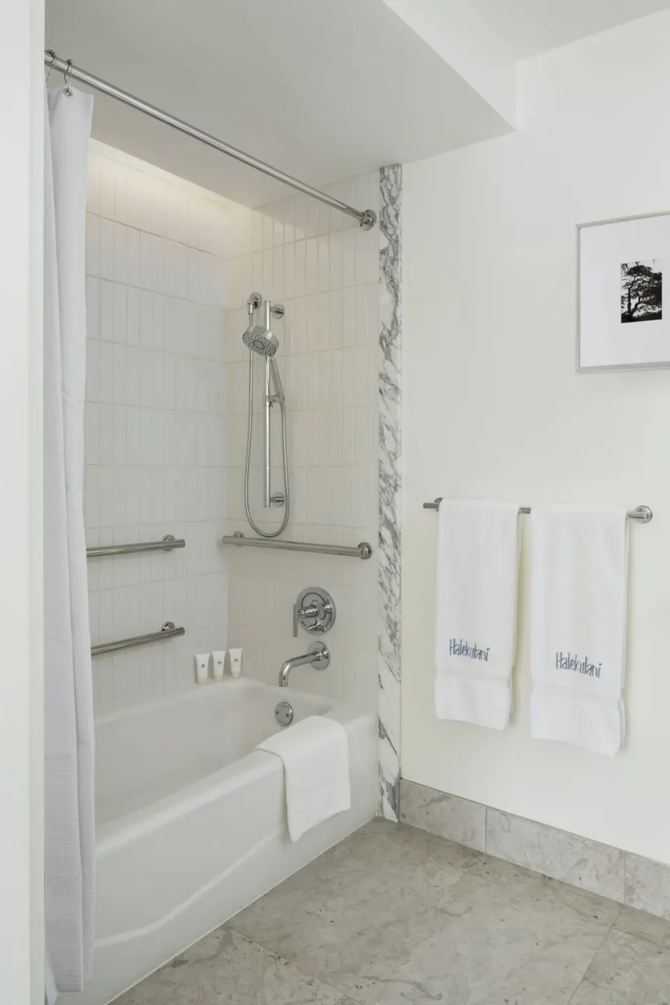 Bathtub and shower with white tile, grab bars, towels, and bath amenities at Halekulani.