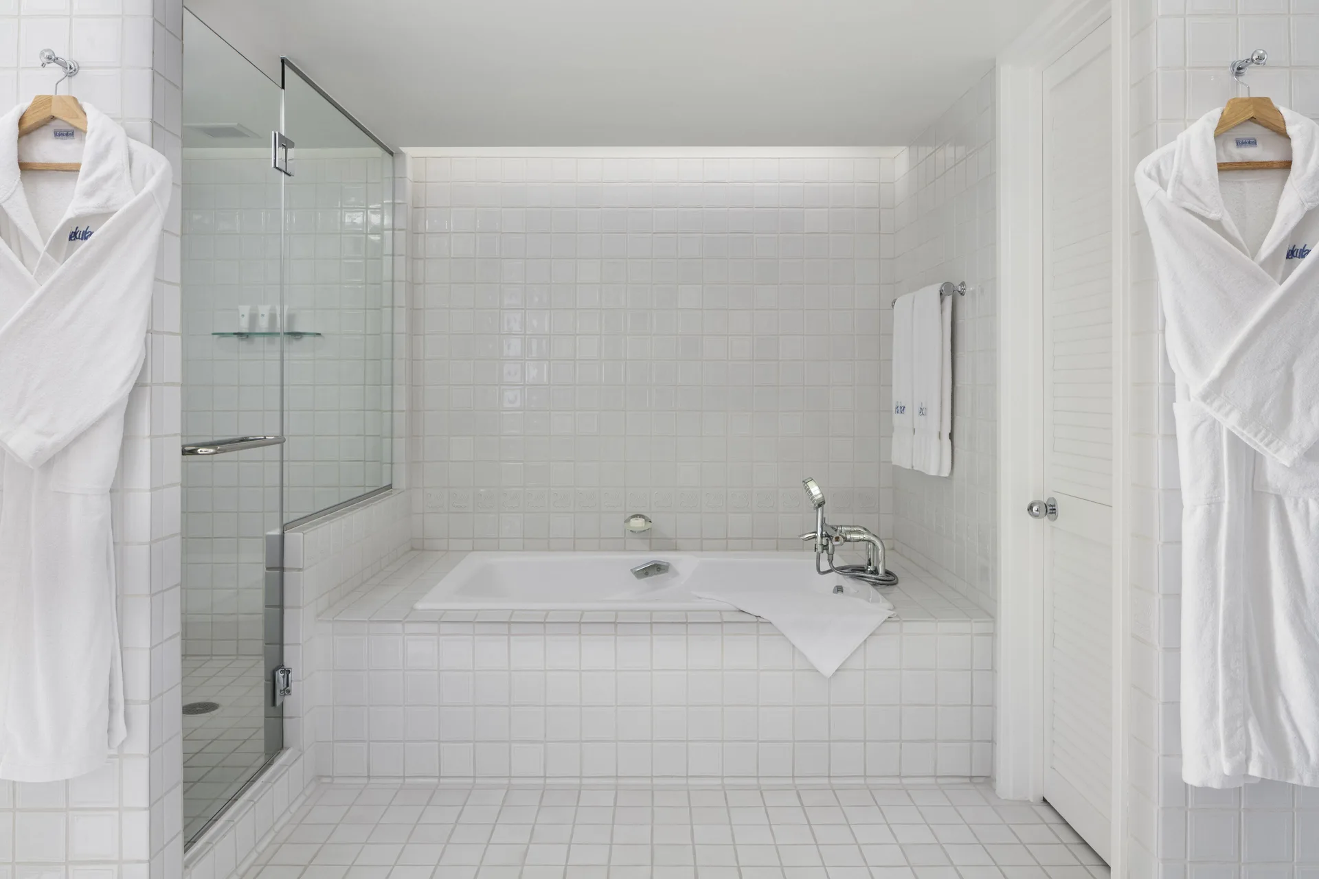 White tiled bathroom with tub, glass shower, robes, and towels at Halekulani.