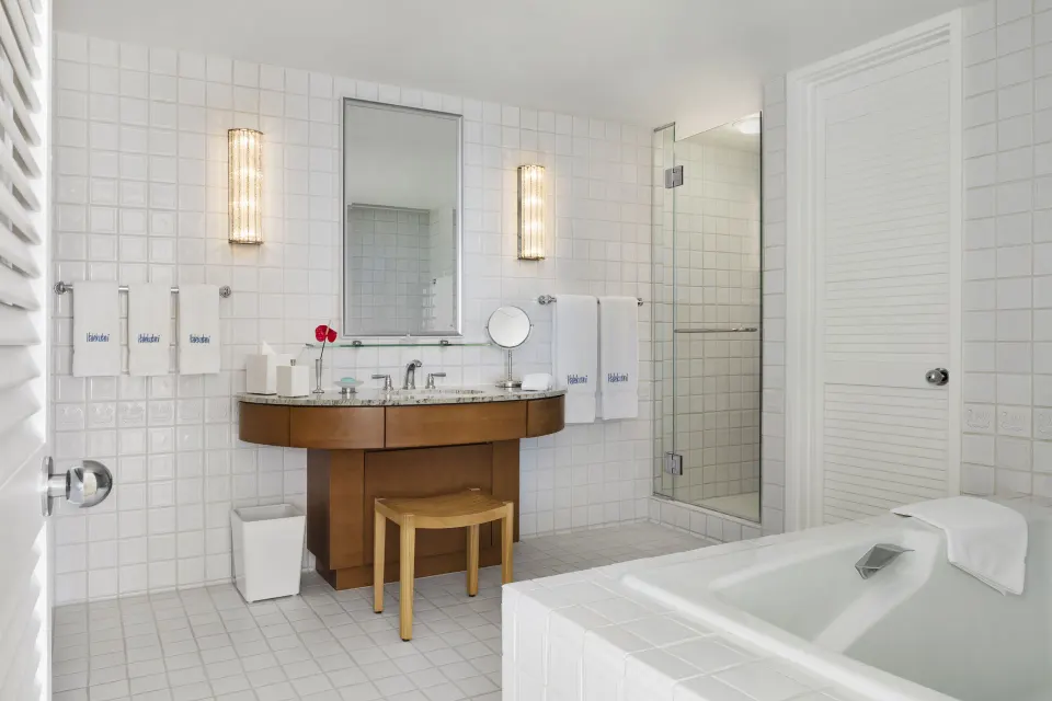 White-tiled bathroom with vanity, tub and glass shower in a Junior Suite at Halekulani.