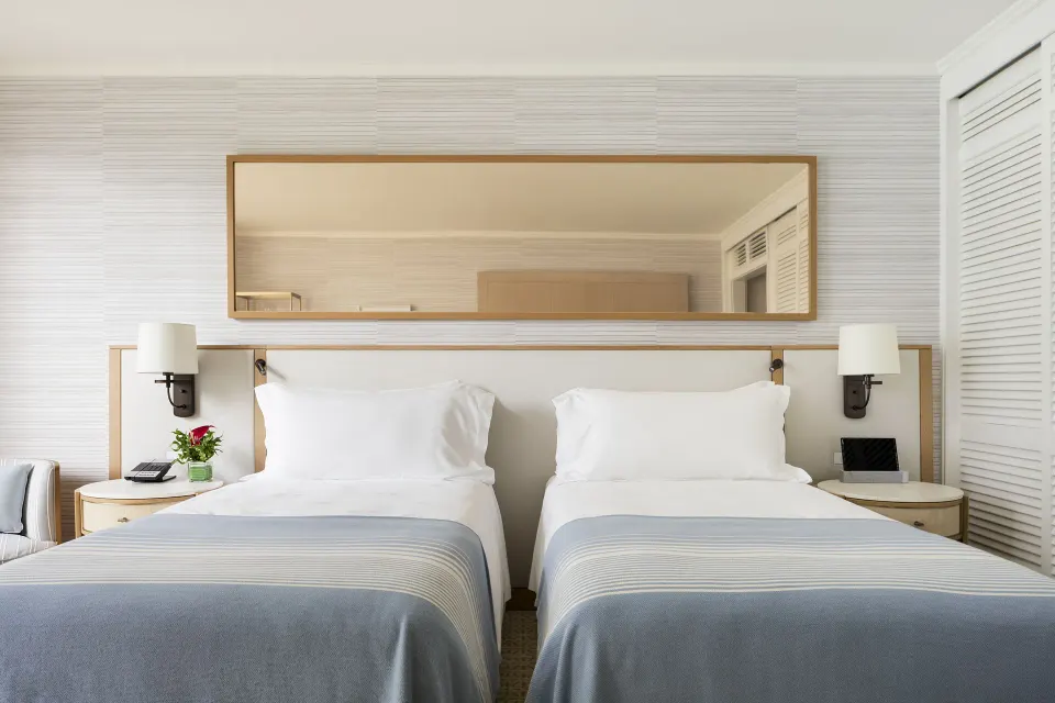 Oceanfront guest room with two beds, wall lamps, mirror, and louvered closet at Halekulani.
