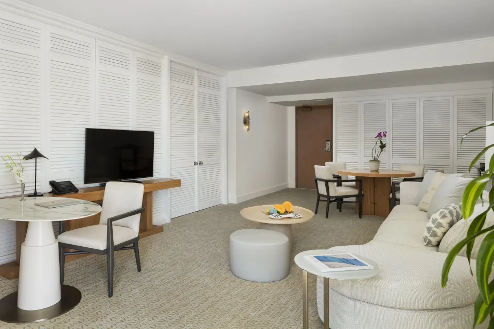 Poolside Suite sitting area with sofa, dining table, TV, and white shutters at Halekulani.