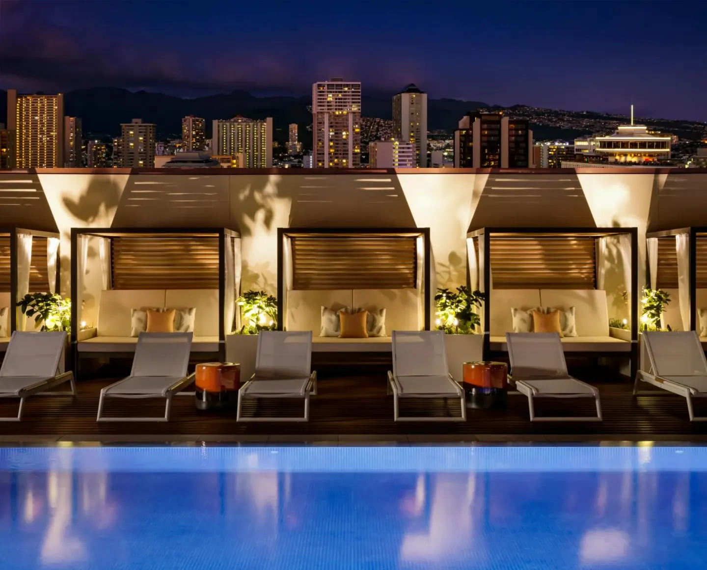 Rooftop pool with loungers, cabanas, and city lights at Halepuna Waikiki by Halekulani.