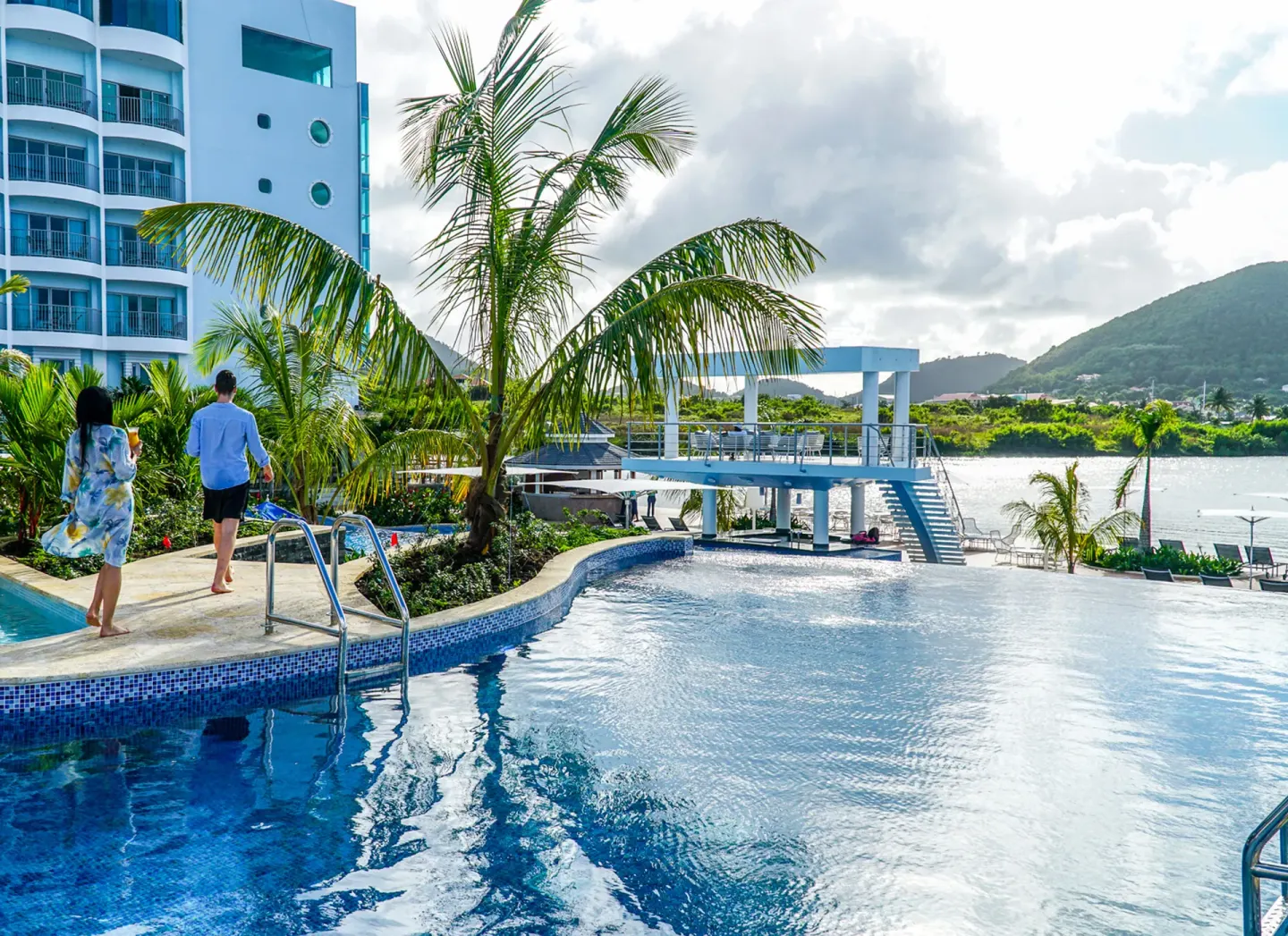 Guests walking beside the pool with palms, lagoon, and resort building at Harbor Club St. Lucia.