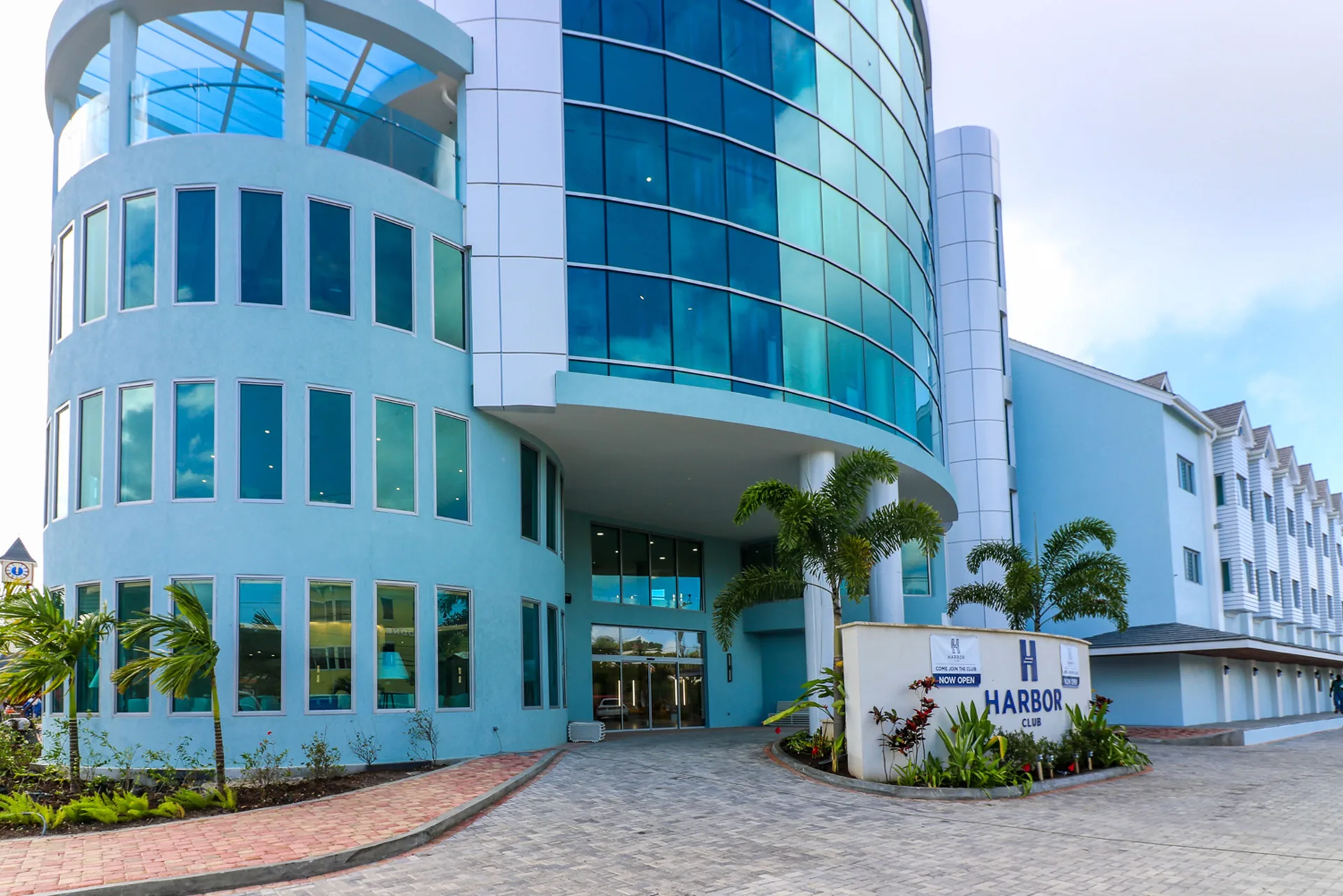 Curved glass-fronted exterior entrance of Harbor Club St. Lucia with palms and signage.