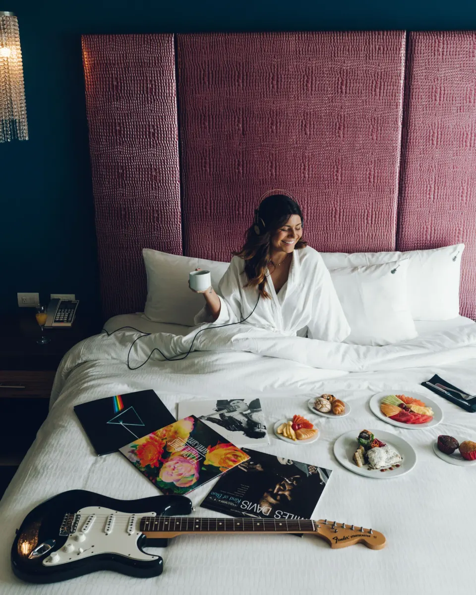 Guest with headphones sits in bed beside breakfast and a guitar at Hard Rock Hotel Cancun.