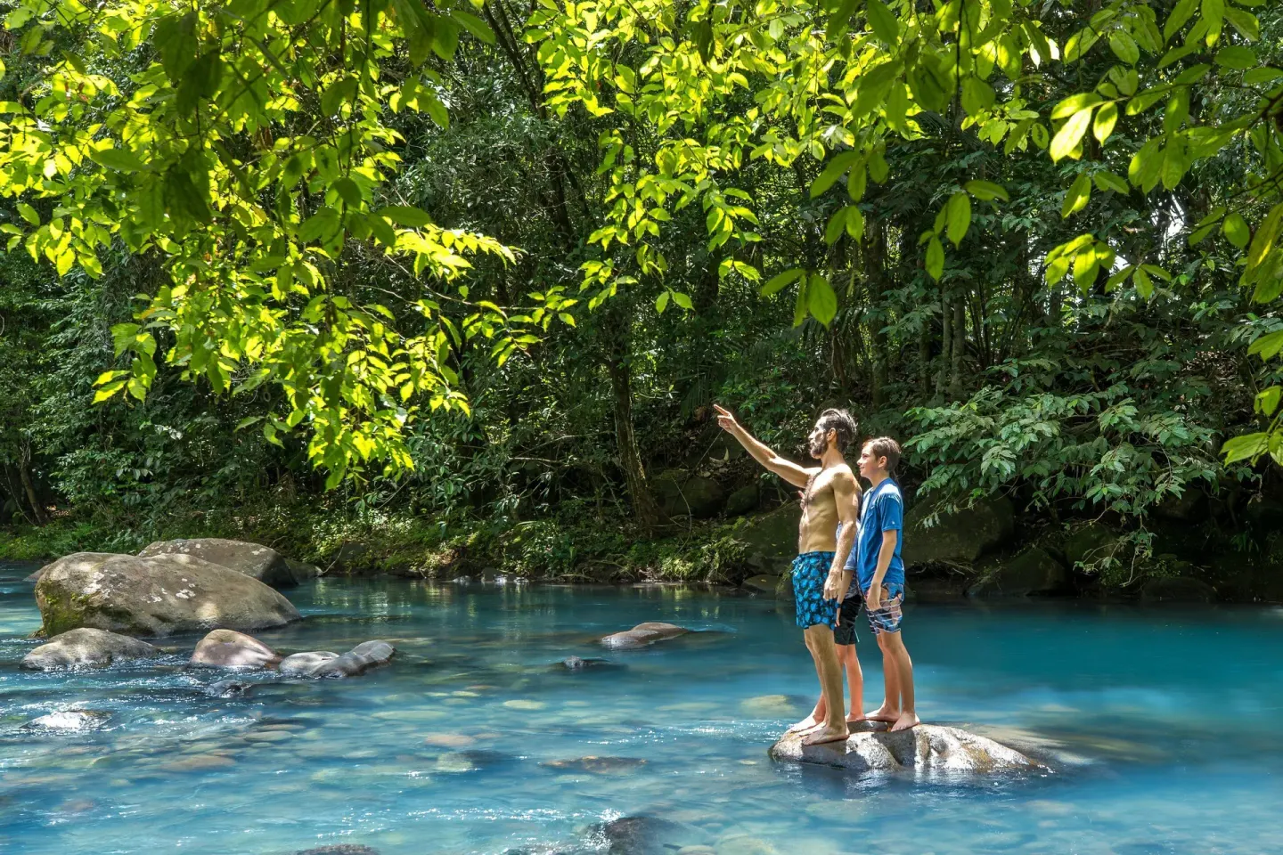 Two guests standing on rocks in a blue river in the rainforest at Hideaway Rio Celeste.