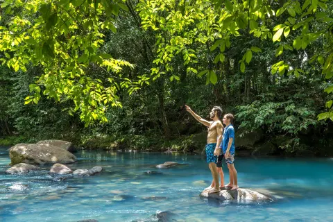 Two guests standing on rocks in a blue river in the rainforest at Hideaway Rio Celeste.
