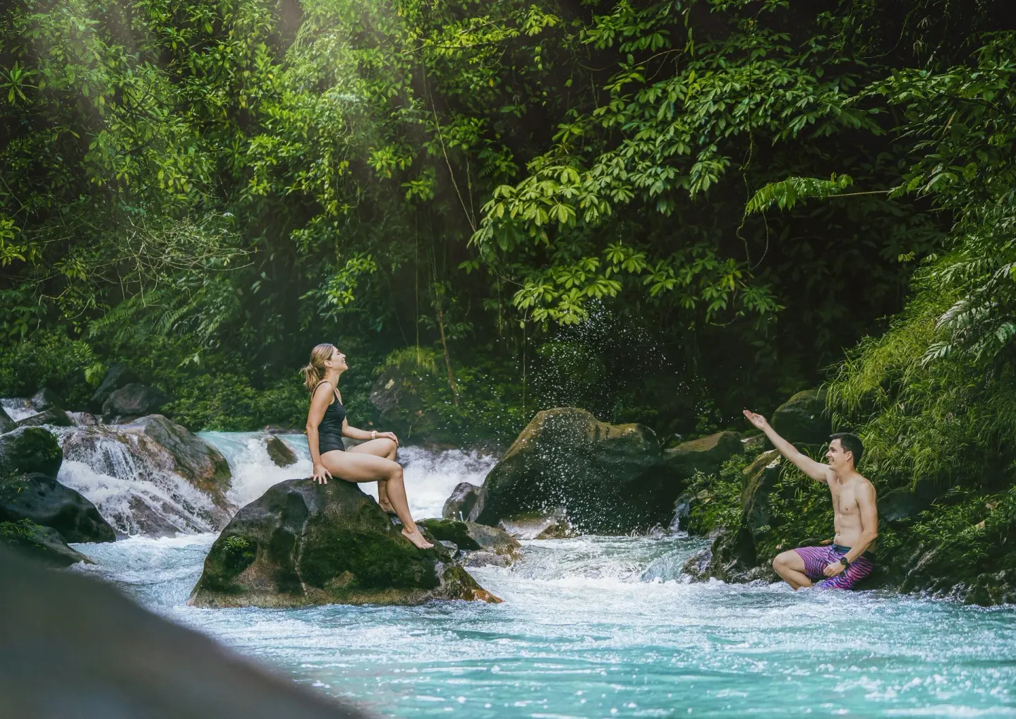Two guests relax by a rocky turquoise river and small rapids at Hideaway Rio Celeste.