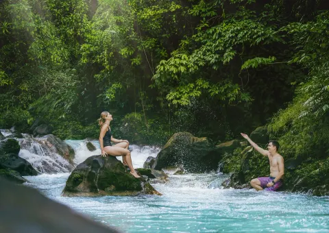 Two guests relax by a rocky turquoise river and small rapids at Hideaway Rio Celeste.