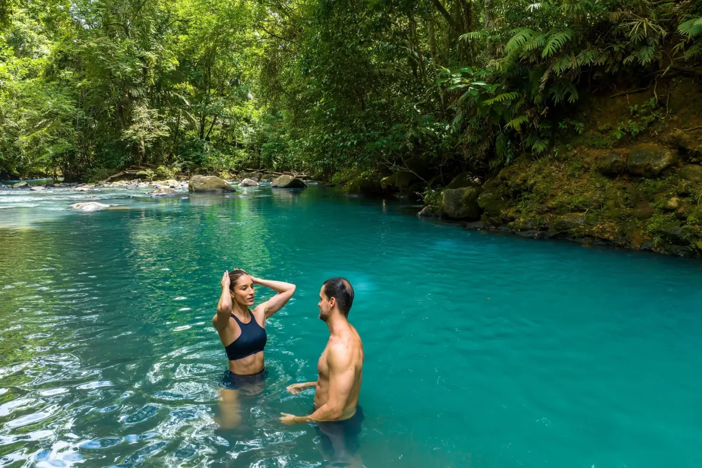 Two guests stand in a clear turquoise river surrounded by forest at Hideaway Rio Celeste.