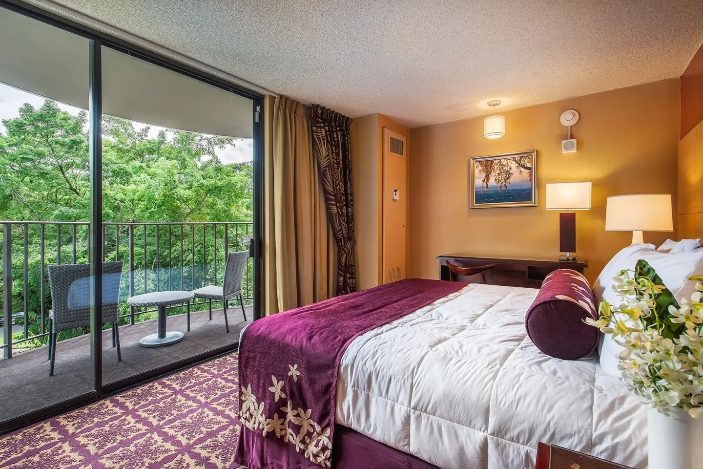 Guest room king bed with balcony seating and tree views at Hilo Hawaiian Hotel.