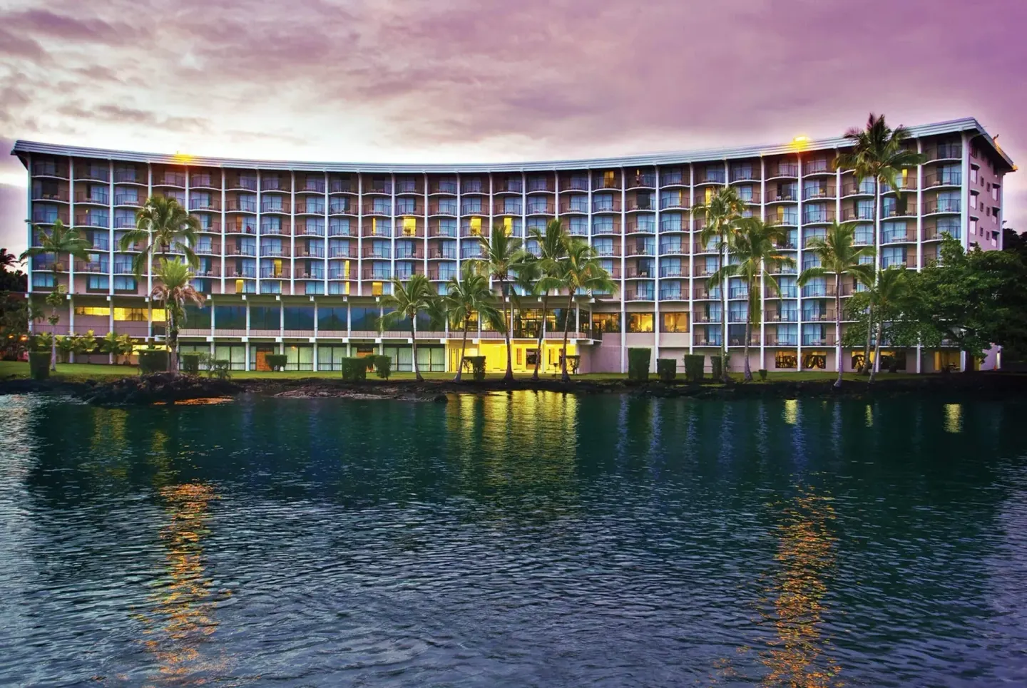 Waterfront exterior of Hilo Hawaiian Hotel with palm trees and balconies at dusk.