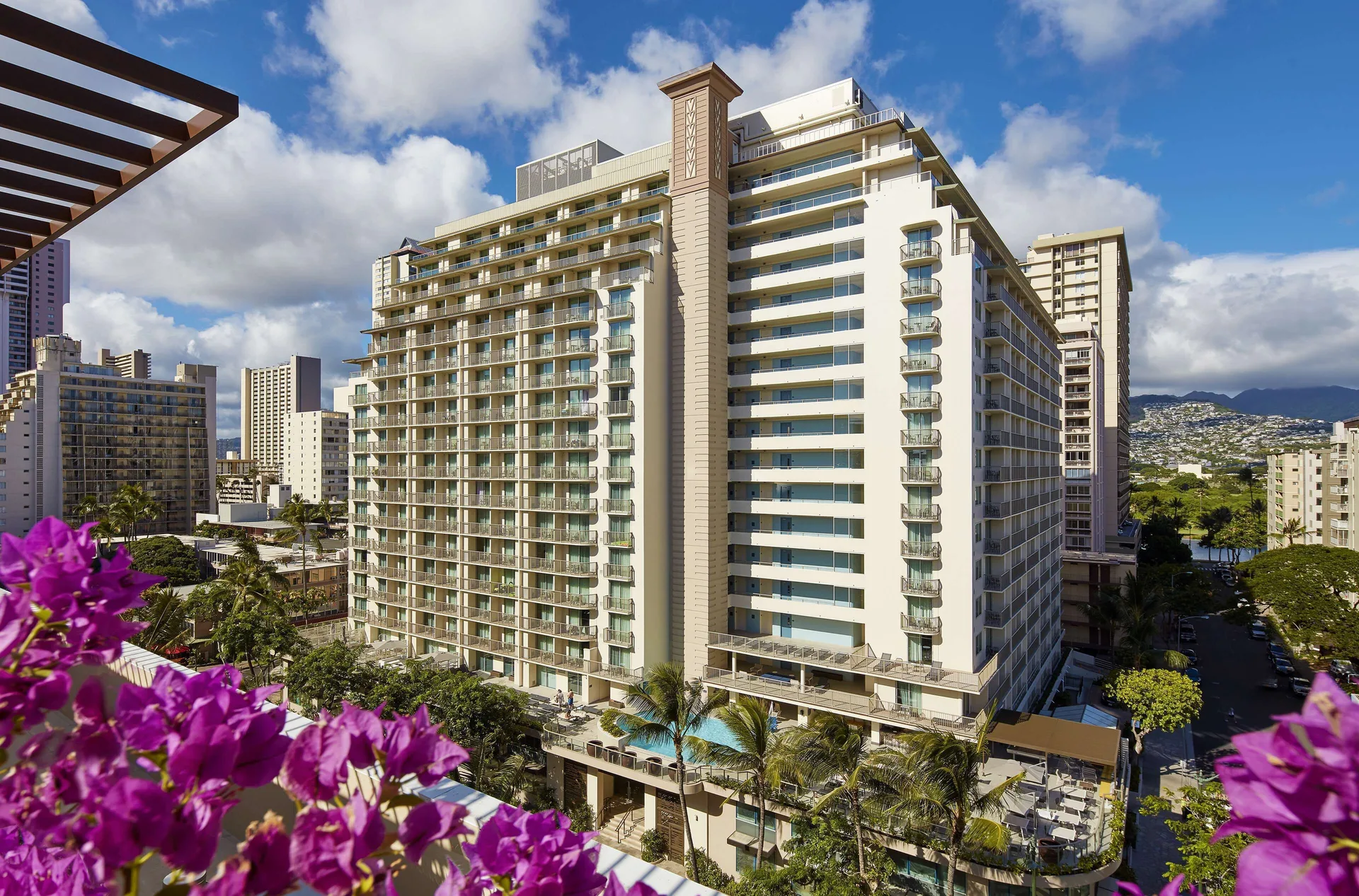 Elevated daytime view of Hilton Garden Inn Waikiki Beach with balconies and pink flowers in front.
