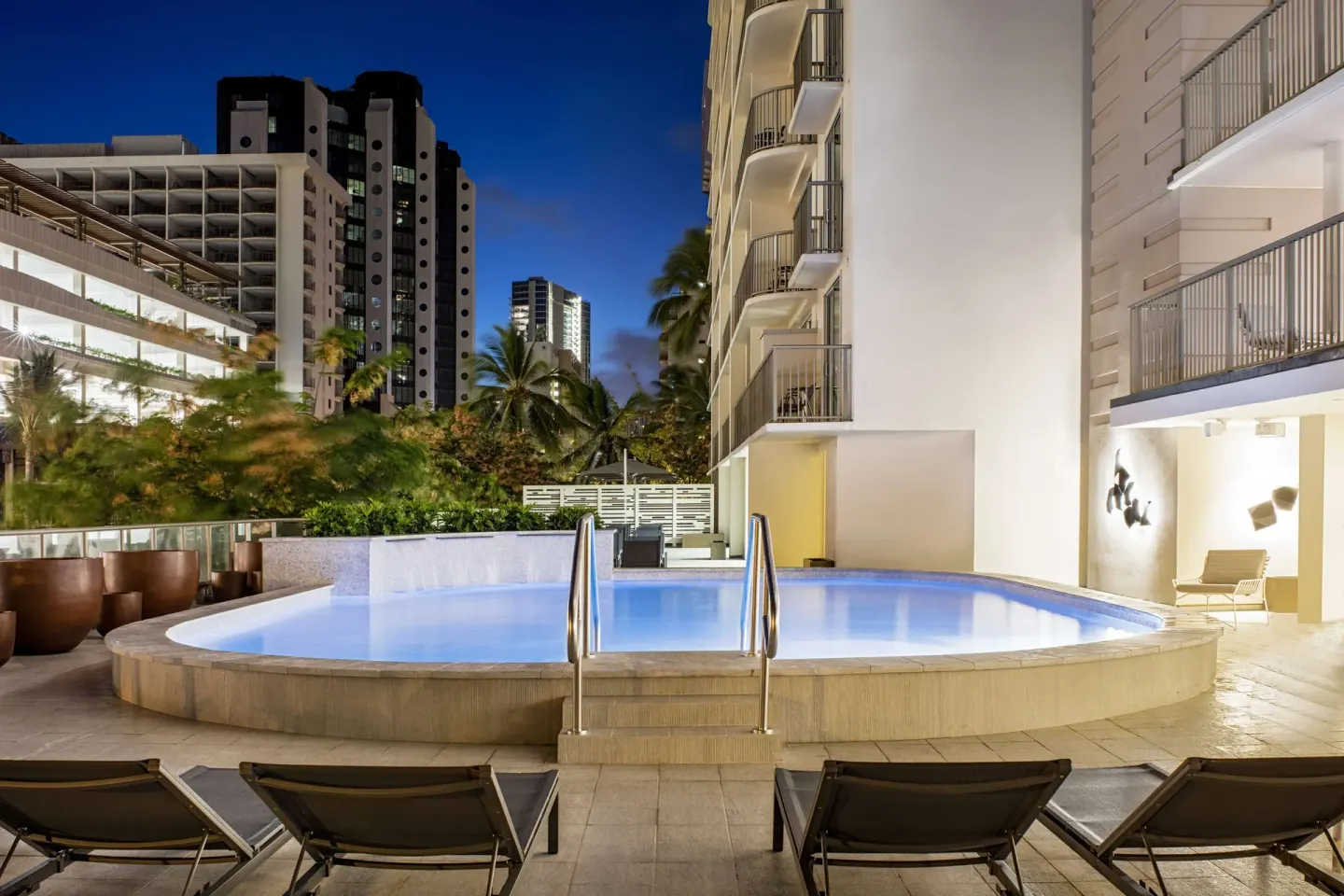 Illuminated evening pool with lounge chairs at Hilton Garden Inn Waikiki Beach.