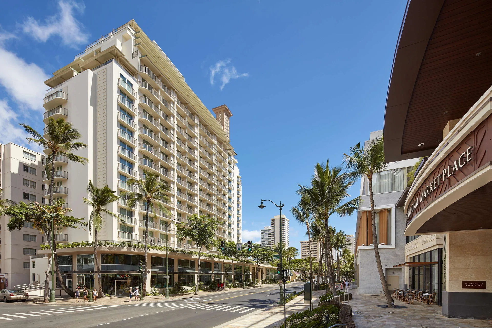 Sunny street view of the Hilton Garden Inn Waikiki Beach tower and palm-lined sidewalk.
