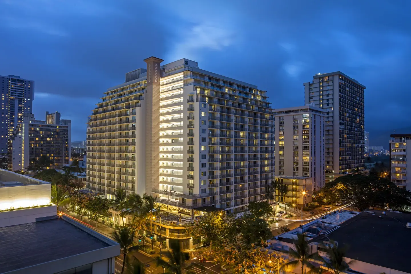 Hotel exterior lit at twilight among Waikiki buildings at Hilton Garden Inn Waikiki Beach.